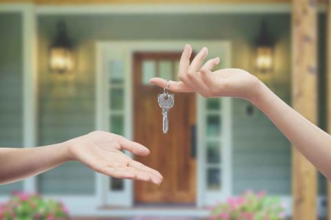 A hand passes keys to another hand in front of a door to signify buying your first home