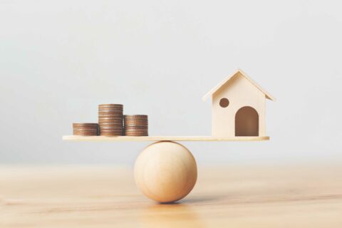 A toy house balances on a wood scale with stacked coins to demonstrate fixed vs variable mortgage decisions