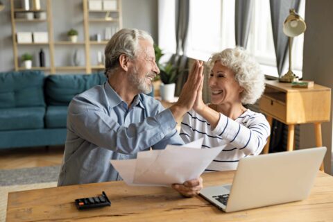 A senior couple high fives in front of a computer, papers, and a calculator to represent the freedom of a reverse mortgage