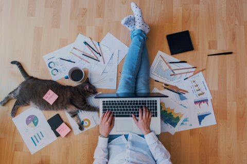 A woman sits on a hardwood floor with a laptop on her legs and charts surrounding her. A cat stretches out beside her and there's a pink sticky note on the cat.